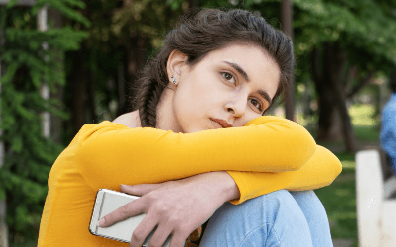 Dark-haired young teen girl in a park, holding a mobile phone and looking sad